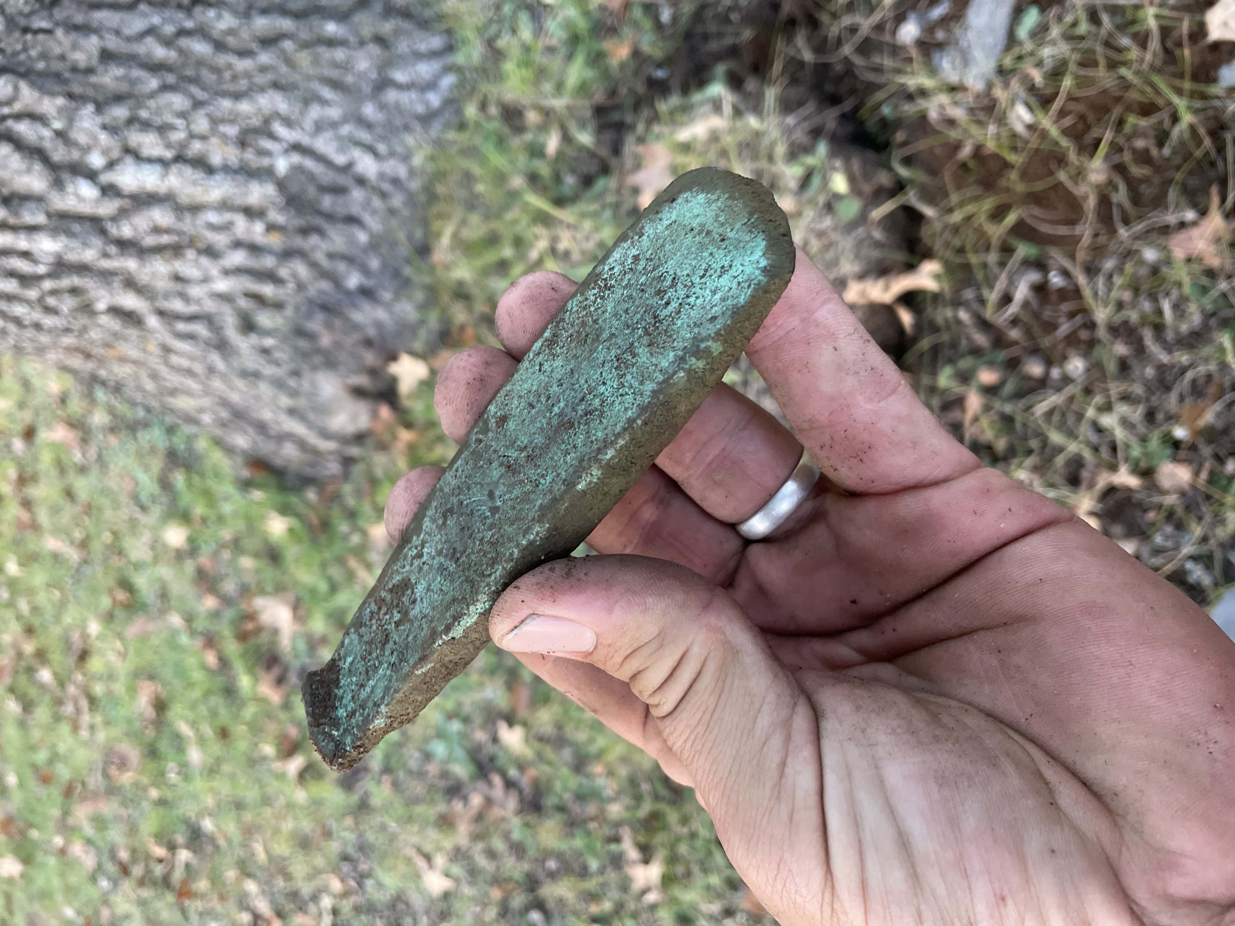 Prehistoric Copper Implement with Green Patina Across Surface Held in a Hand with Grassy Park and Tree Blurry in Background