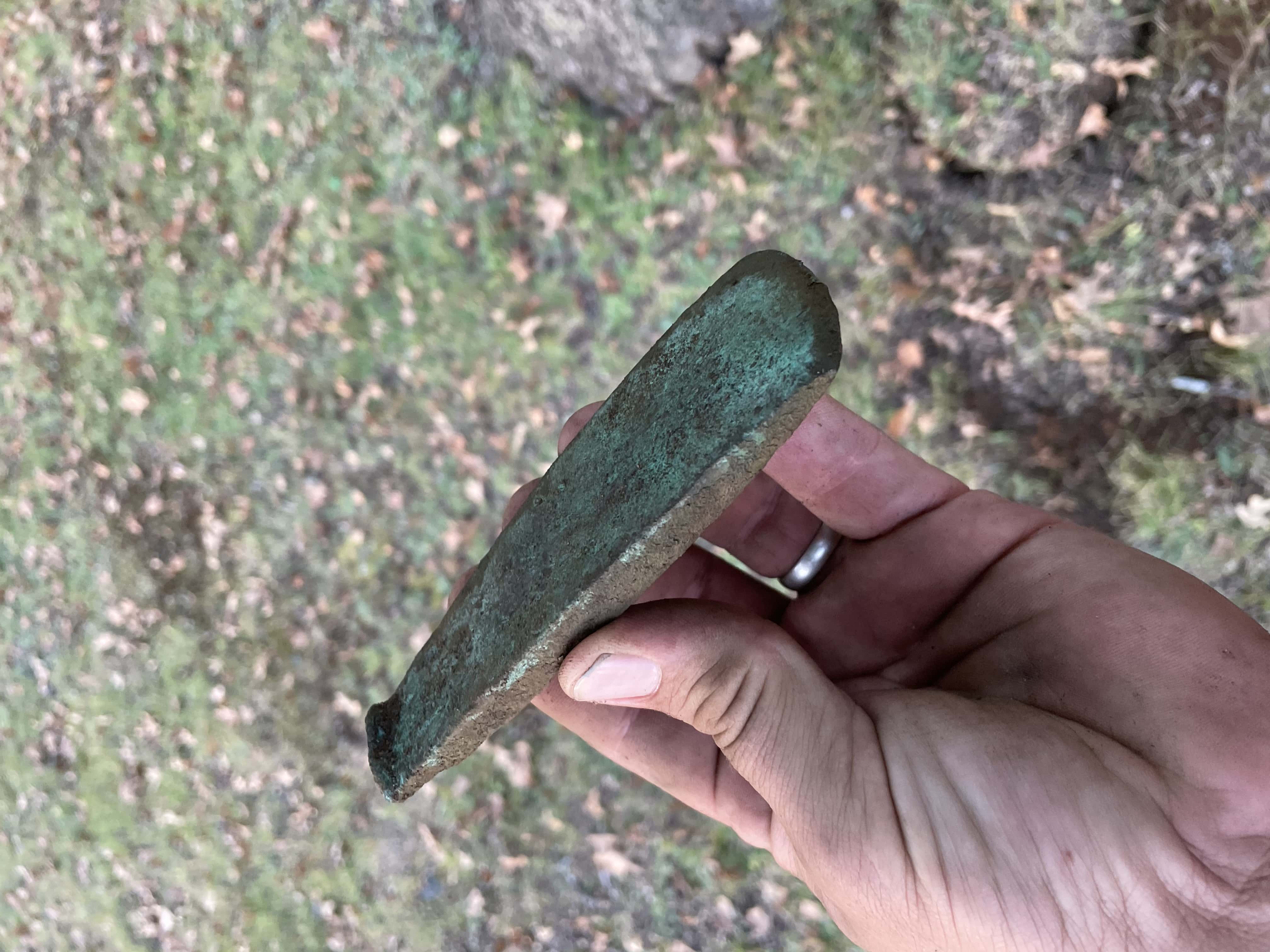 Prehistoric Copper Implement with Green Patina Across Surface Held in a Hand with Grassy Park and Tree Blurry in Background