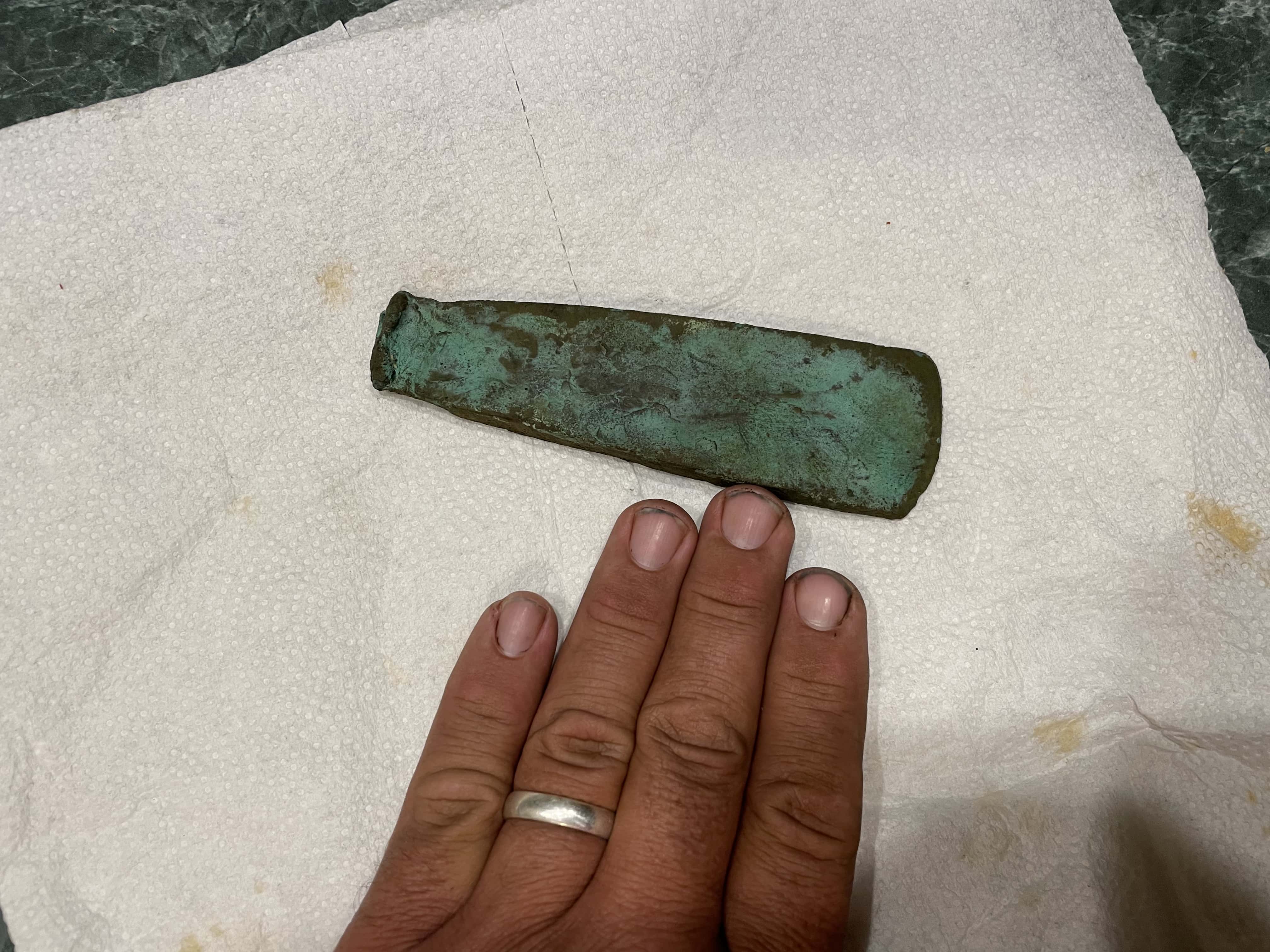 Prehistoric Copper Implement with Green Patina Across Surface Sat on Paper Towel with Hand Laid Flat Beneath it to Show the Size