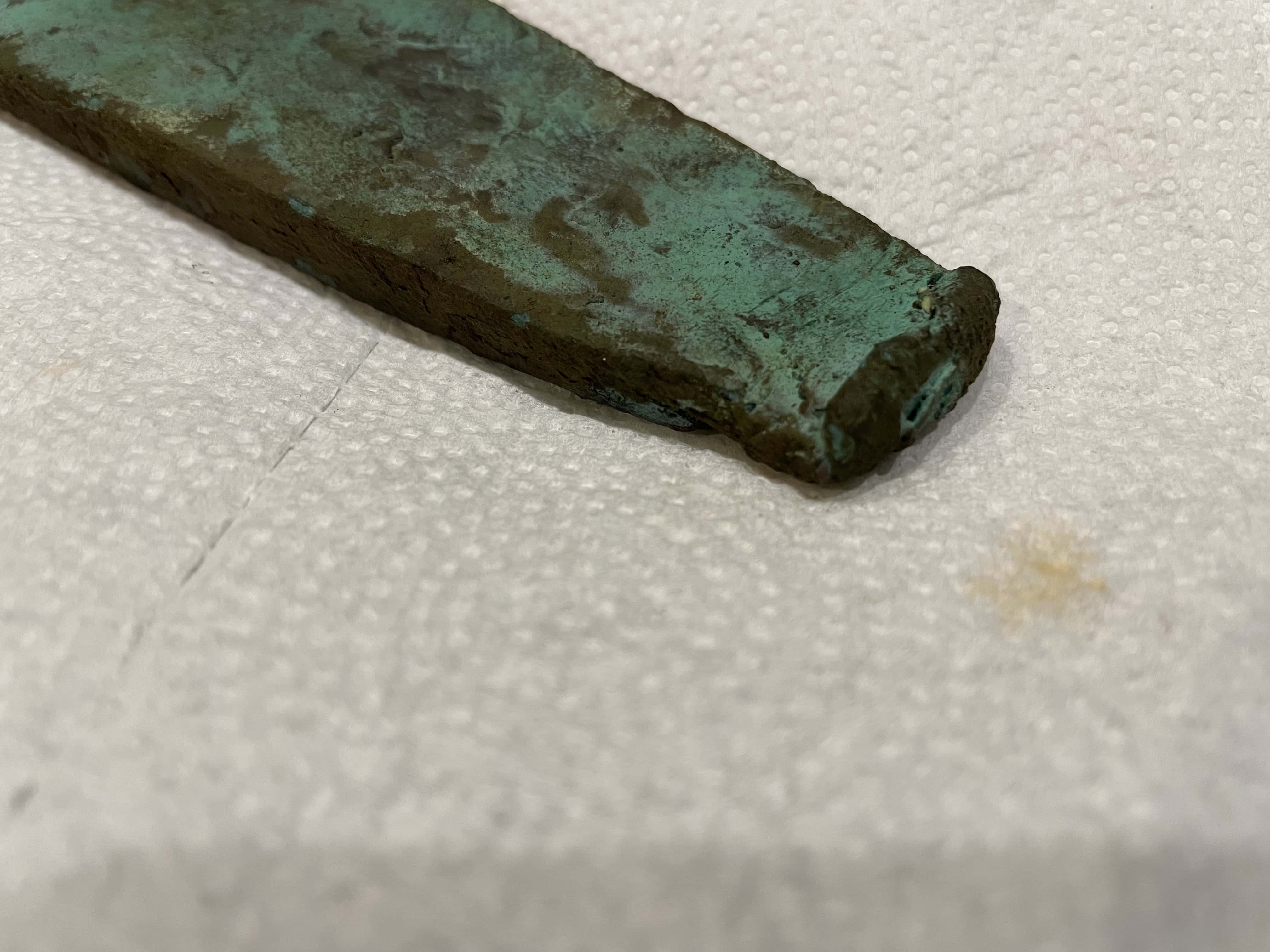 Prehistoric Copper Implement with Green Patina Across Surface Showing End Edge Resting On Paper Towel in Background
