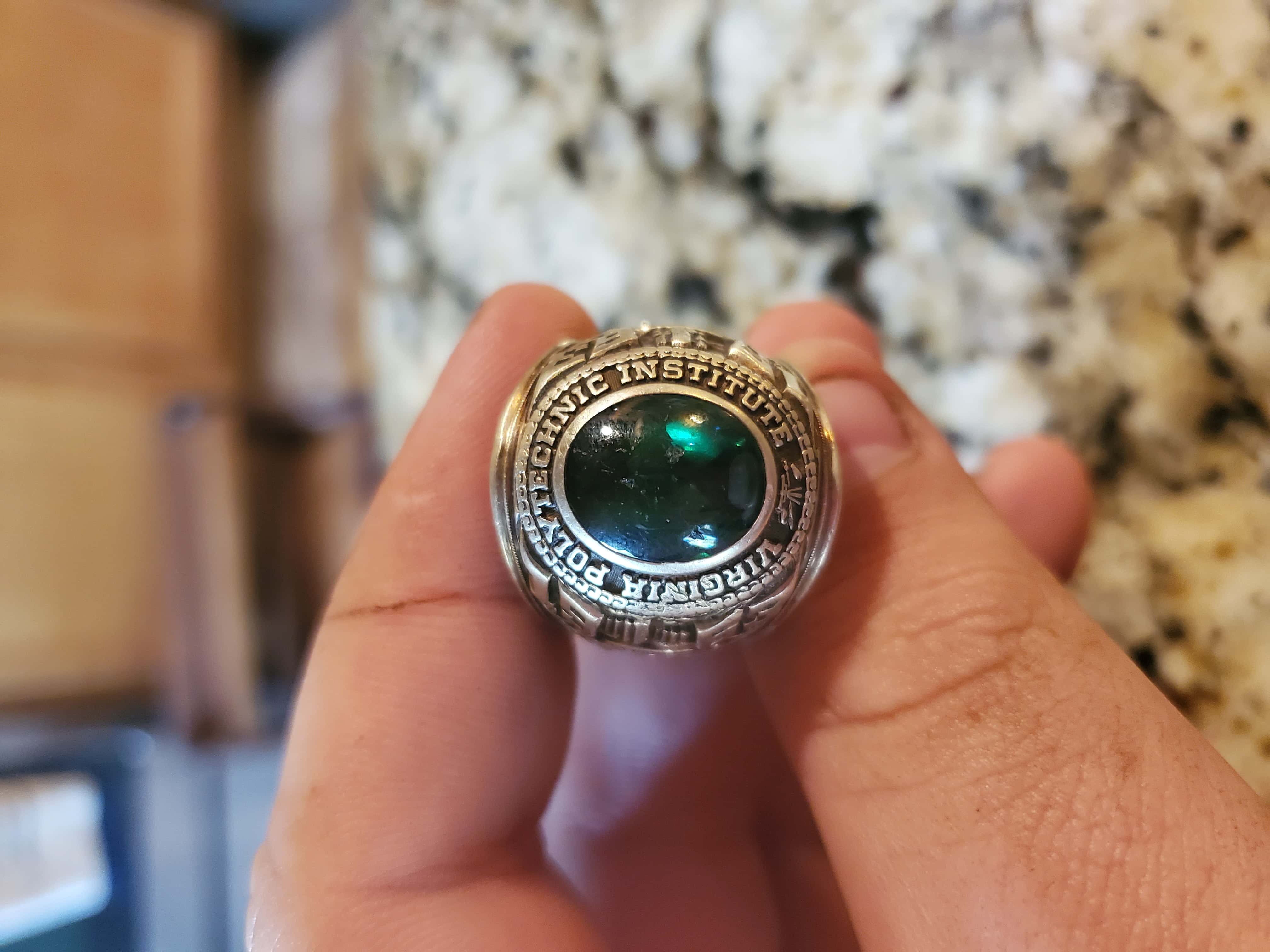Image of Ring Held Between Forefinger and Thumb with Beautiful Green Colored Gemstone or Emerald at Center and Virginia Polytechnic Institute Around the Edge with Other Details Visible at Edge and Bezel